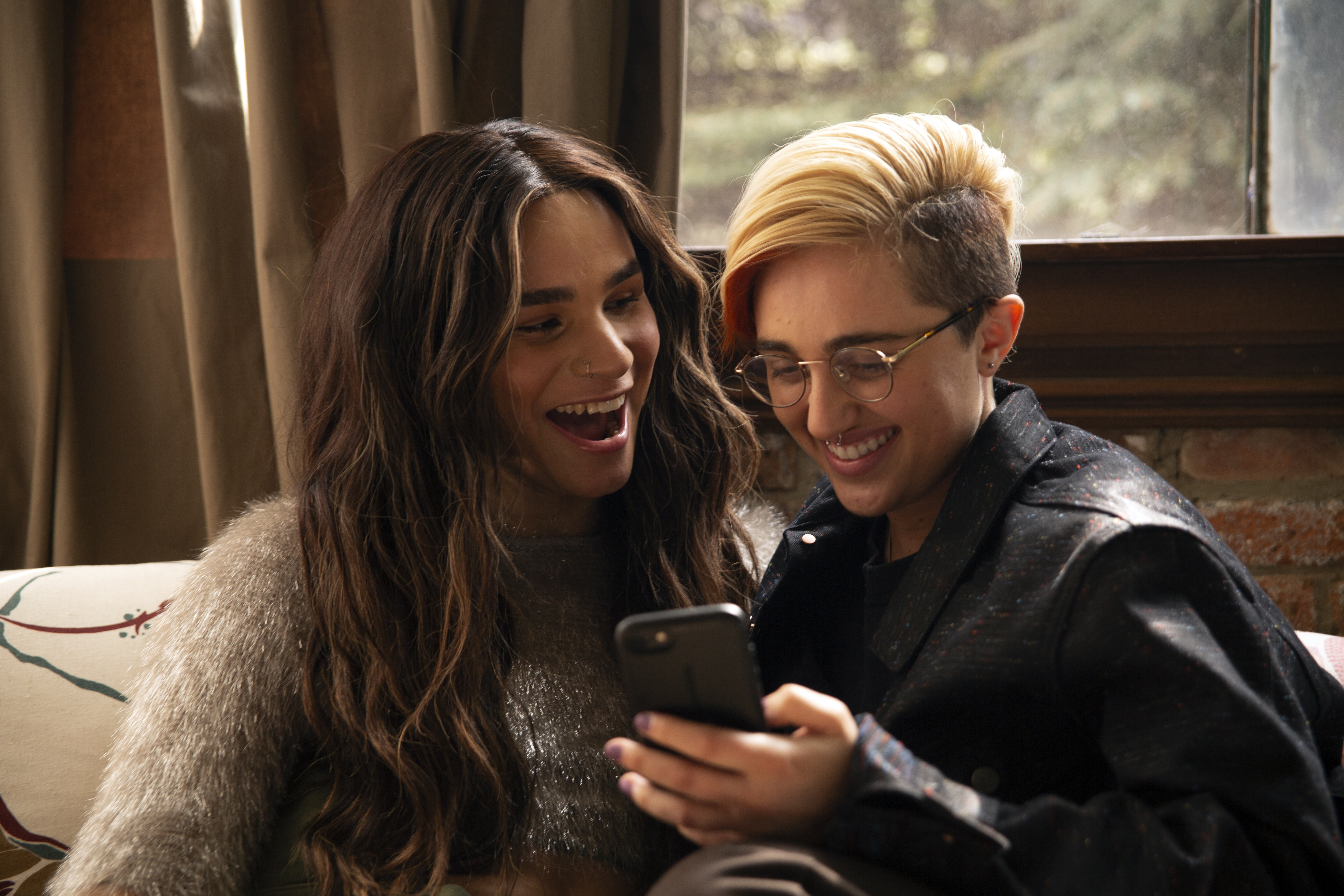 A transfeminine non-binary person and transmasculine gender-nonconforming person laughing and looking at a phone