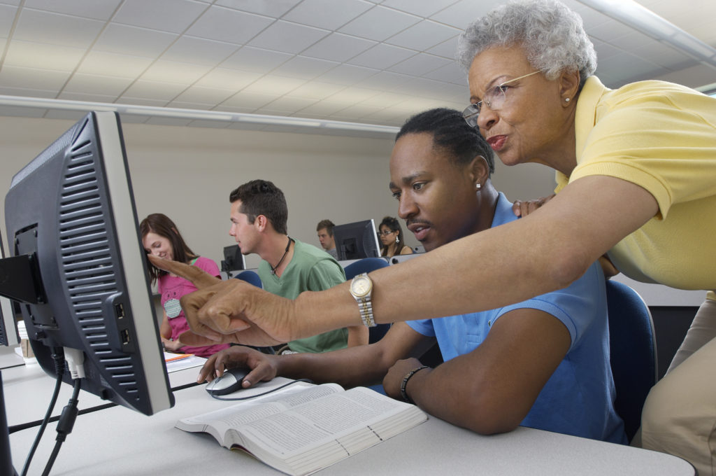 Senior teacher assisting male student during computer class with classmates in the background