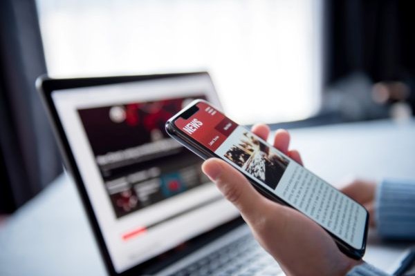 Mindful Media Habits: Photo of hand holding smartphone with news headlines on screen and laptop in background.