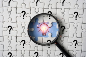 A magnifying glass focused on a lightbul in the middle of puzzle pieces with question marks on them
